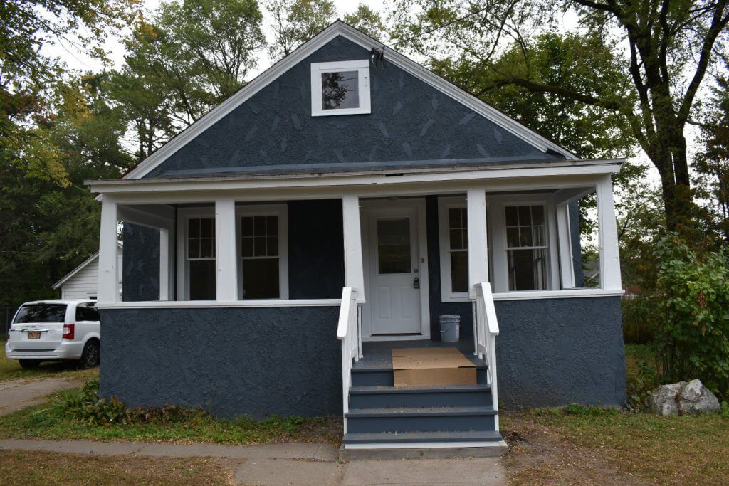 A beautifully updated blue stucco home in Glens Falls, NY, featuring white trim, a welcoming covered porch, and fresh exterior paint