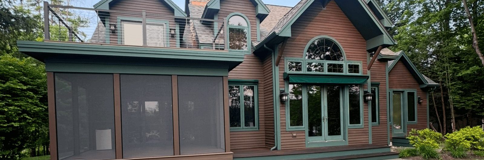 Cabin staining project showcasing a two-story wood home with freshly stained brown siding, green trim accents, screened porch, and upper balcony for a rustic yet elegant finish