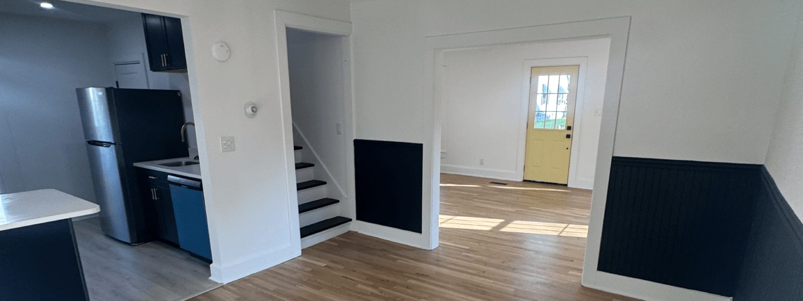 "Interior painting project featuring a modern open layout with freshly painted white walls, deep navy wainscoting, refinished hardwood floors, updated kitchen with stainless steel appliances, and a bright yellow entry door