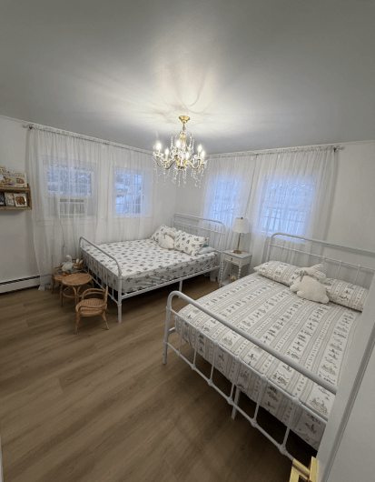 Renovated children’s bedroom featuring matching twin beds, soft neutral decor, natural light from multiple windows, and hardwood-style floors