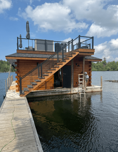 An exterior view of a freshly stained brown log boathouse situated on a calm lake. A wooden pier leads to the structure, and the side docks are covered in protective drop cloths. The boathouse features a black metal staircase leading up to a rooftop deck with matching railings. On the right side, a person is standing on a step ladder working on the boathouse wall under a partly cloudy blue sky.
